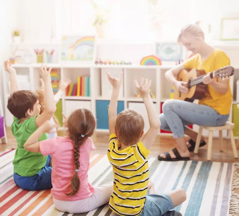 osoba grająca na gitarze i dzieci siedzące na dywanie